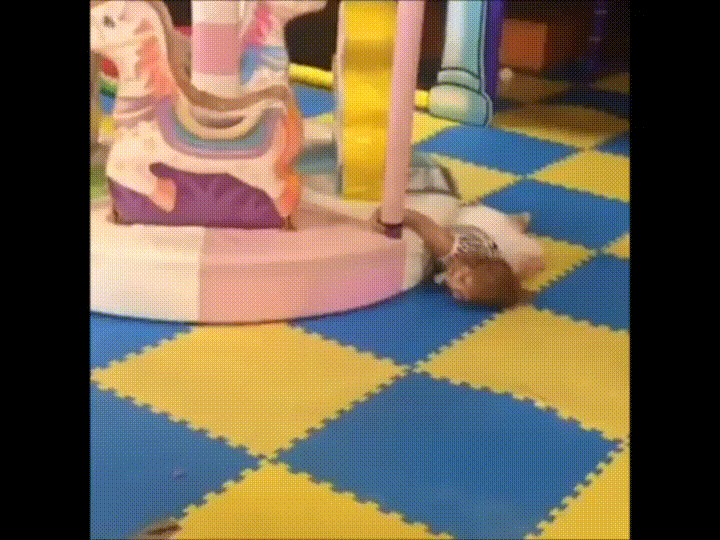 animated gif of a young girl holding onto a merry-go-round with a single hand. She is laying face down on the floor and the merry-go-round is dragging her along the floor as it spins, slowly, in an infinite animated gif loop.